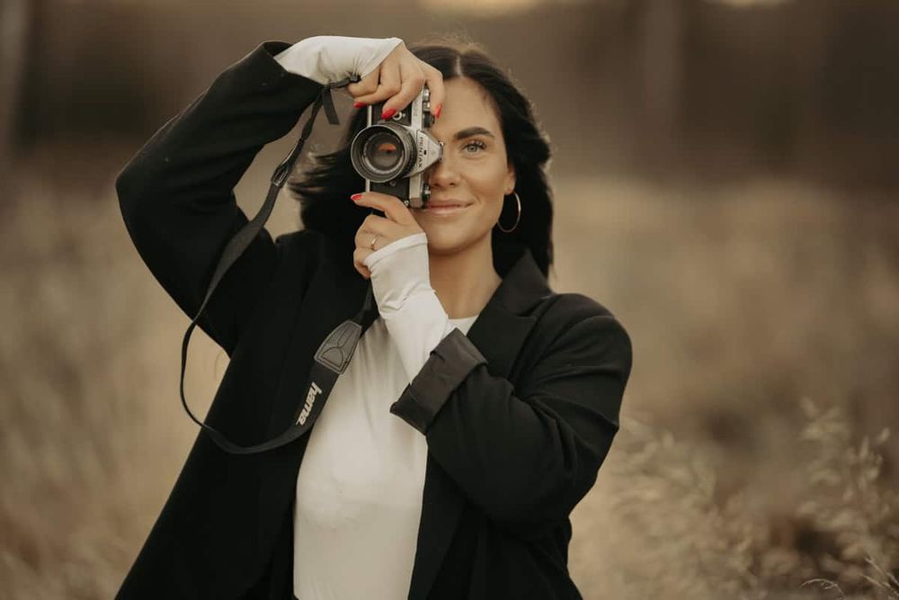 Person holding vintage camera while wearing black coat and white top in outdoor moody setting.