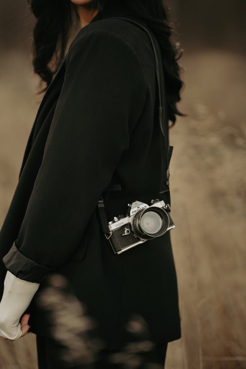 Person wearing black blazer carries vintage film camera on strap against moody brown background in artistic portrait.