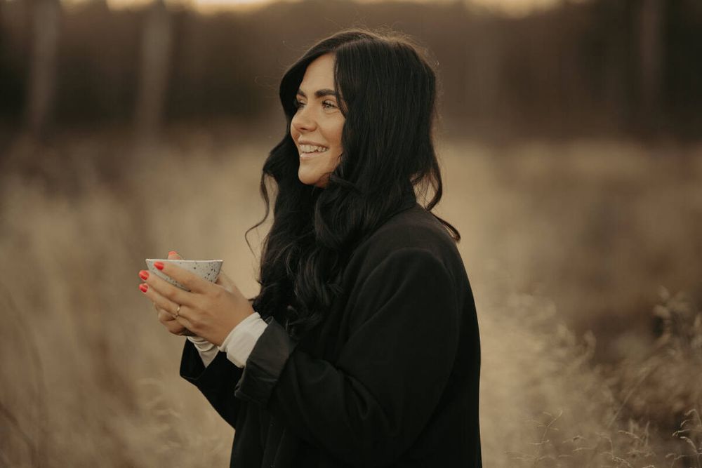 Someone with long dark hair wearing a black jacket holds a cup while smiling outdoors in a golden-hour field setting.