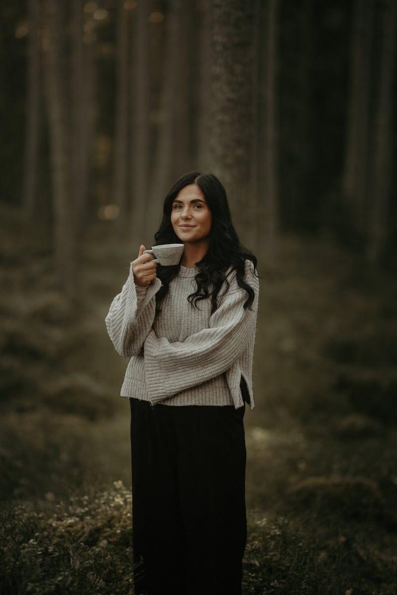 Someone wearing a cozy knit sweater and long black skirt stands in a misty autumn forest holding a warm drink.