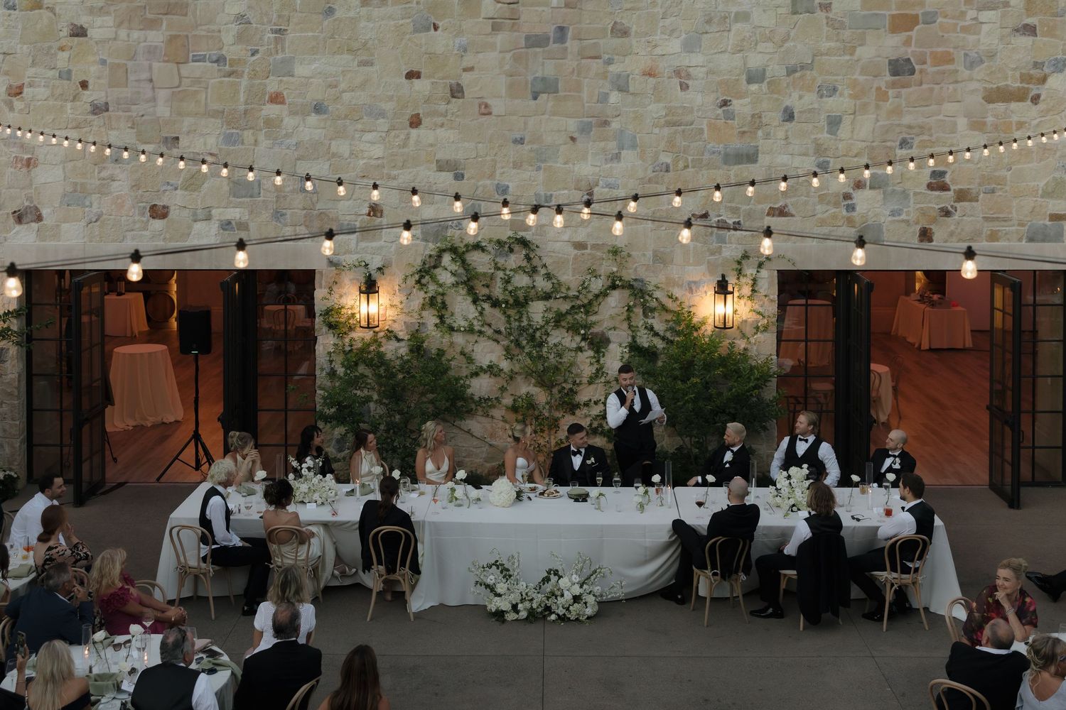 A couple shares their first kiss during an elegant outdoor garden wedding ceremony surrounded by white floral arrangements.