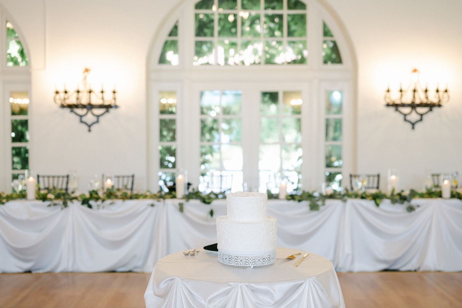 Elegant white wedding cake stands centered in front of draped tables at an airy venue with arched windows and wall sconces.