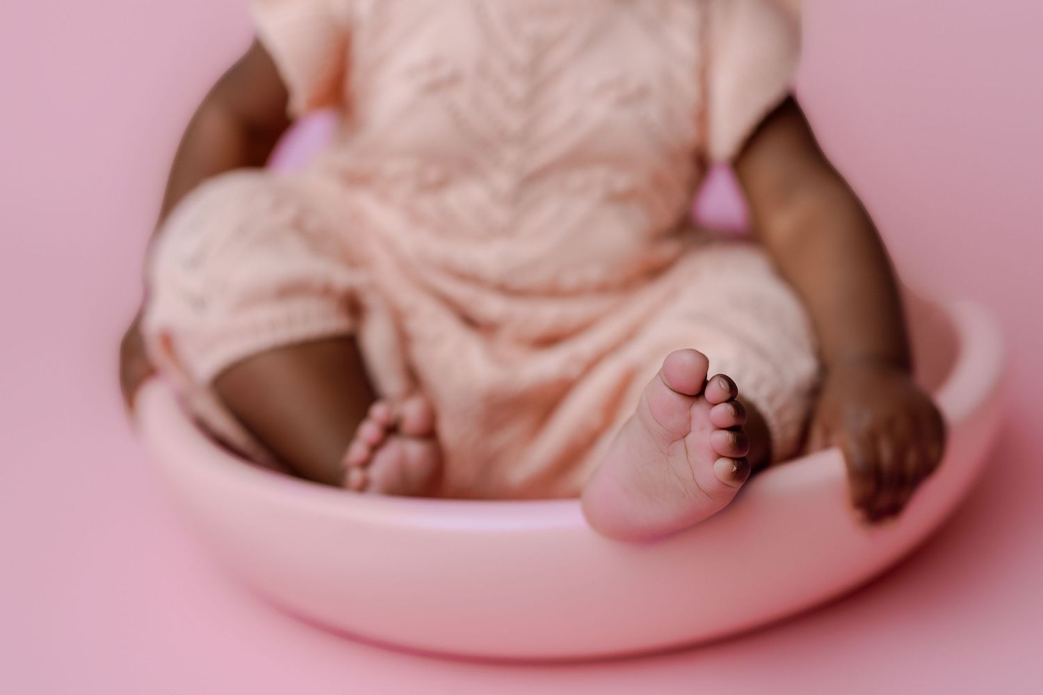 Close up of tiny infant feet and legs wearing a peachy pink outfit against a soft pink background.