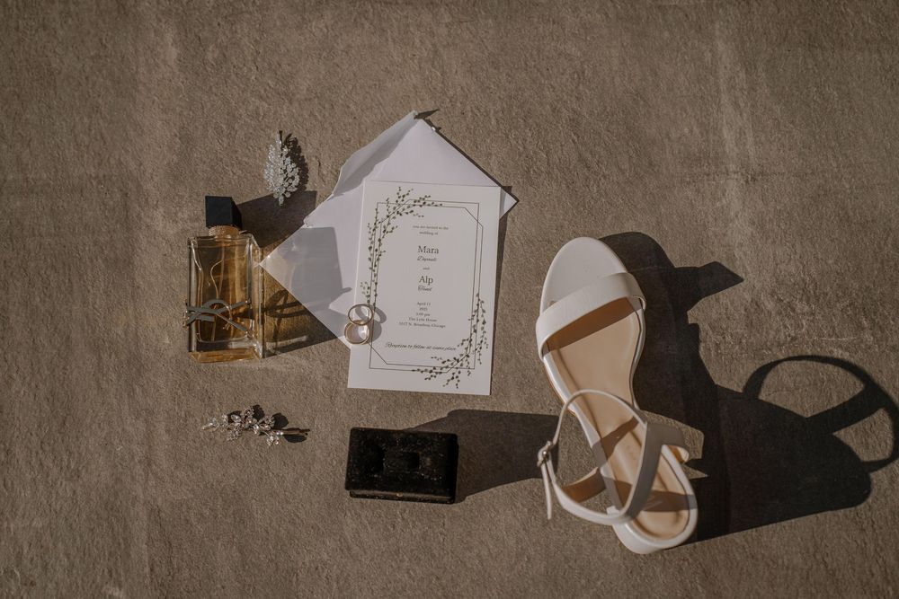 Wedding accessories including invitation card, sandals, and phone arranged on beige textured surface with dramatic shadows.