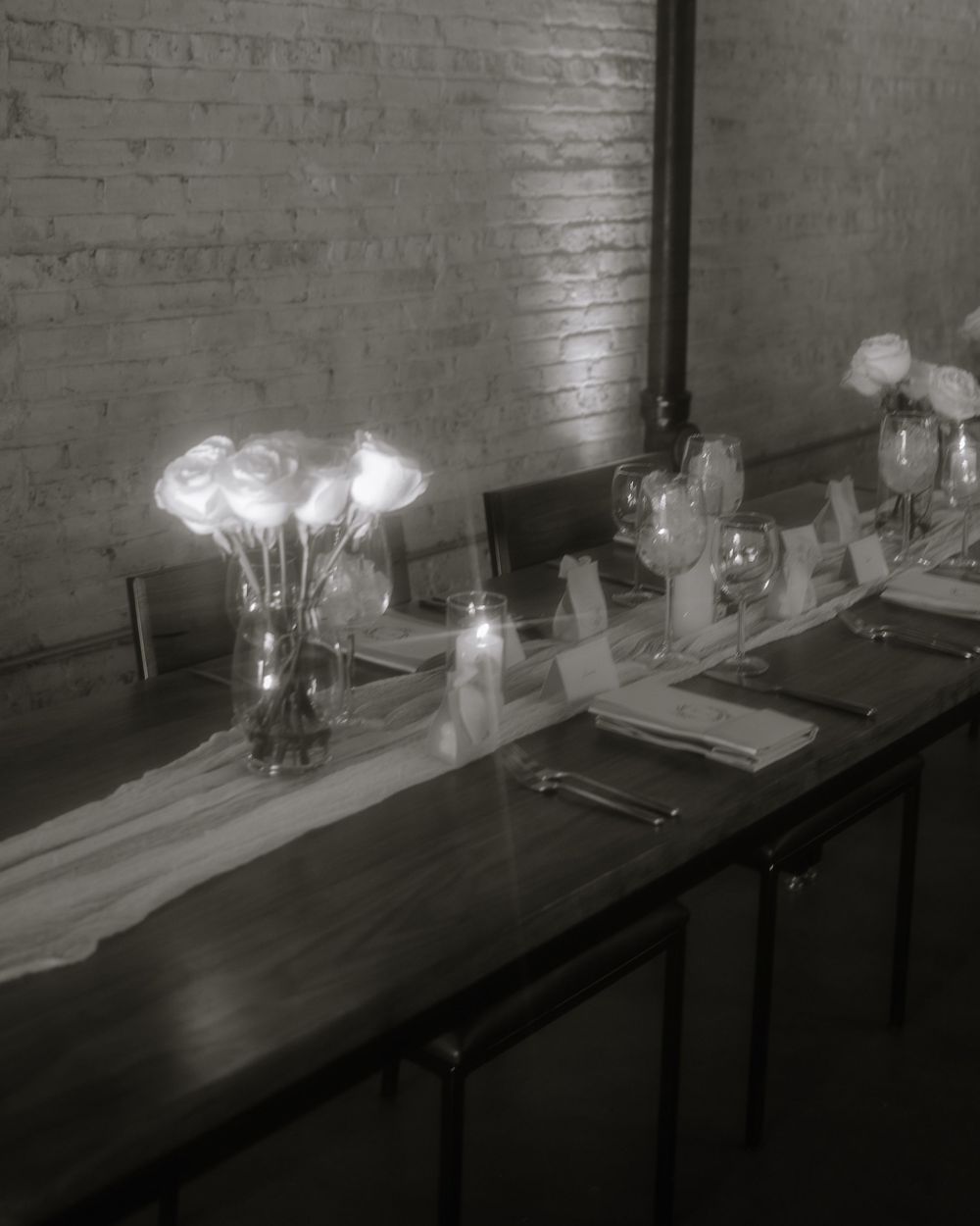 An elegant black and white table setting with glass vases of white flowers and place settings against a brick wall.