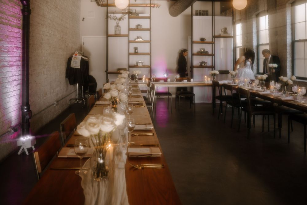 Long wooden dining table set for an elegant wedding reception with candlelight in an industrial-style loft space.
