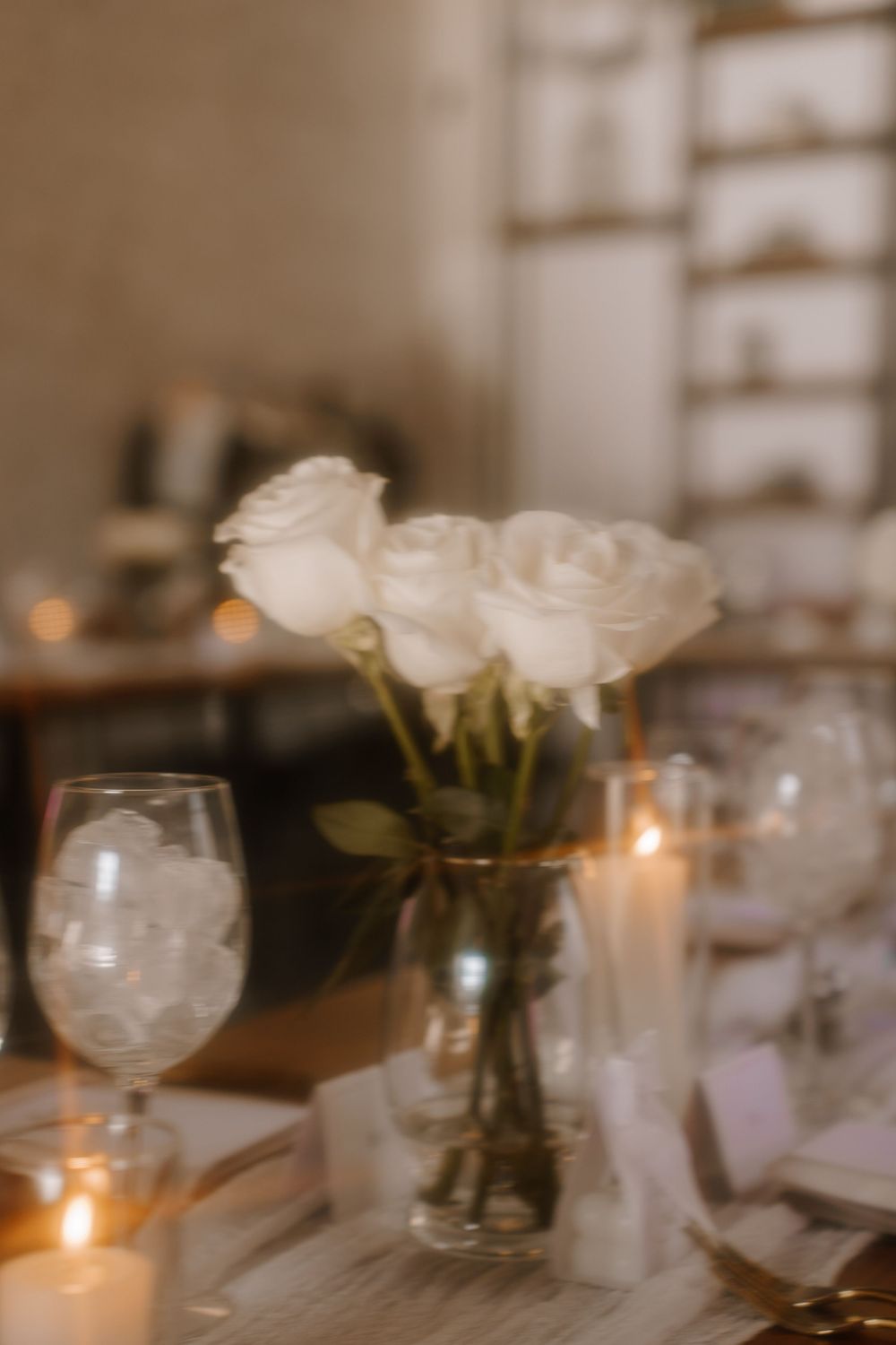 Elegant table setting with white roses in vase, lit candles and glassware creating romantic restaurant ambiance.