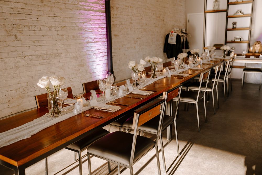 A long wooden dining table set with white linens and place settings against exposed brick walls in a modern restaurant space.