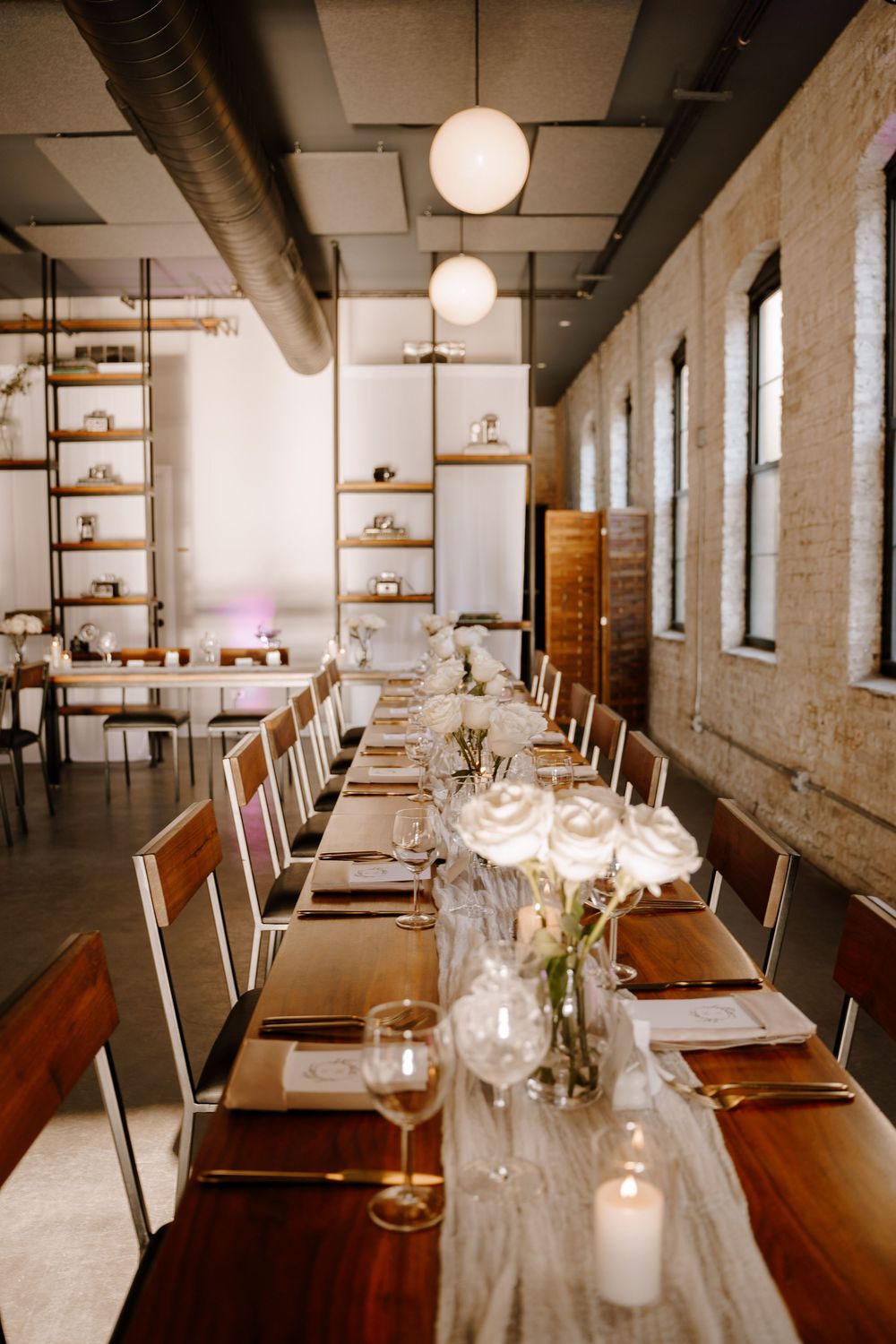 An elegant table setting with glowing candles and white floral centerpieces in an industrial-style dining room with pendant lights.