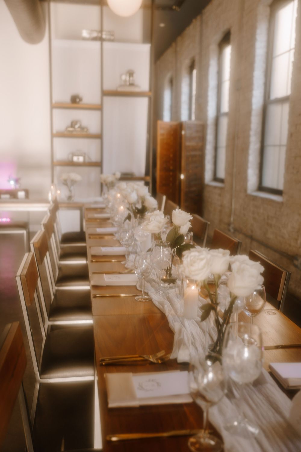 A rustic wedding reception table decorated with white floral centerpieces and wooden chairs in a brick-walled venue.