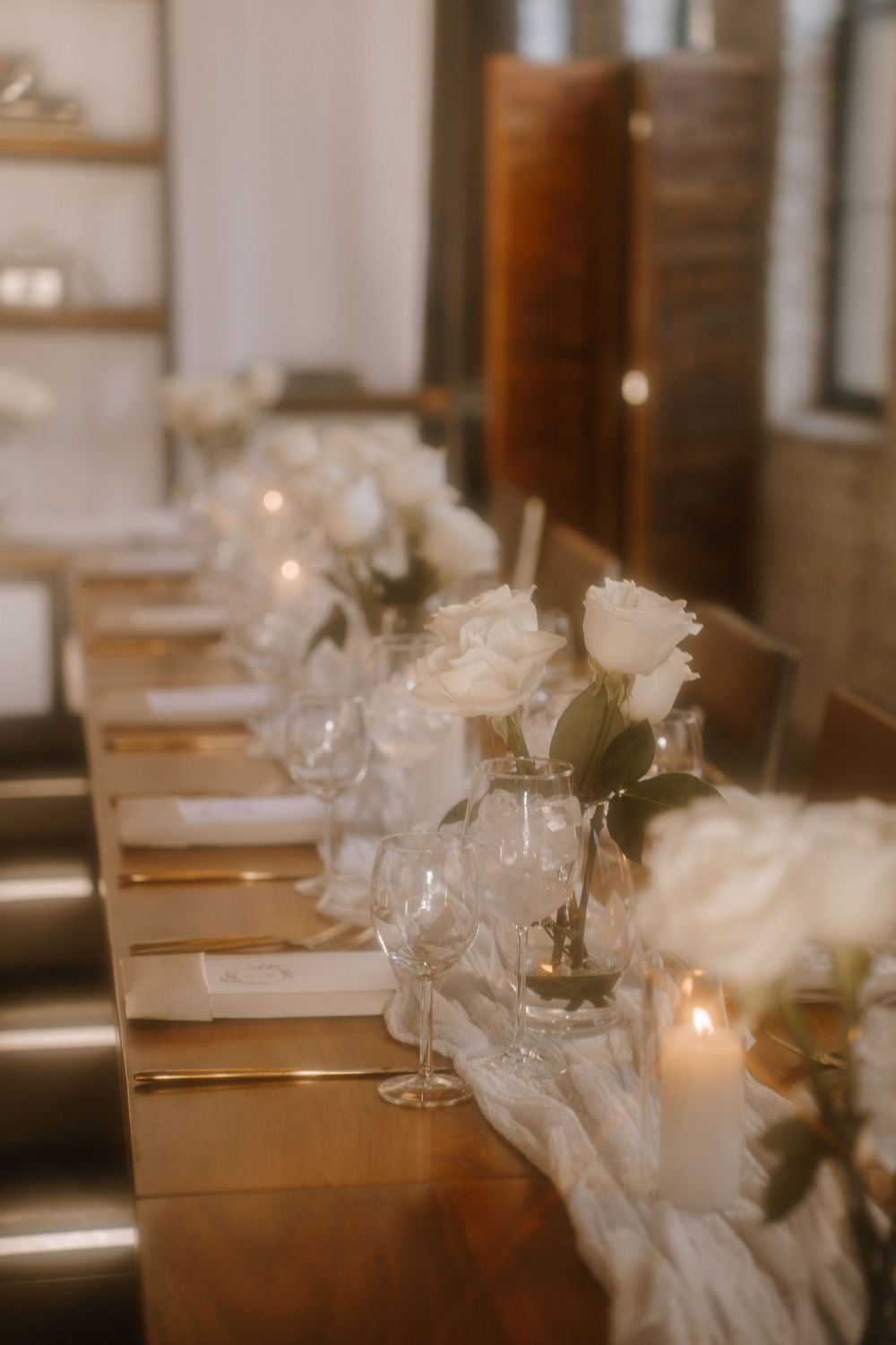 Elegant wedding table setting with white roses and candles along a wooden dining table in warm lighting.