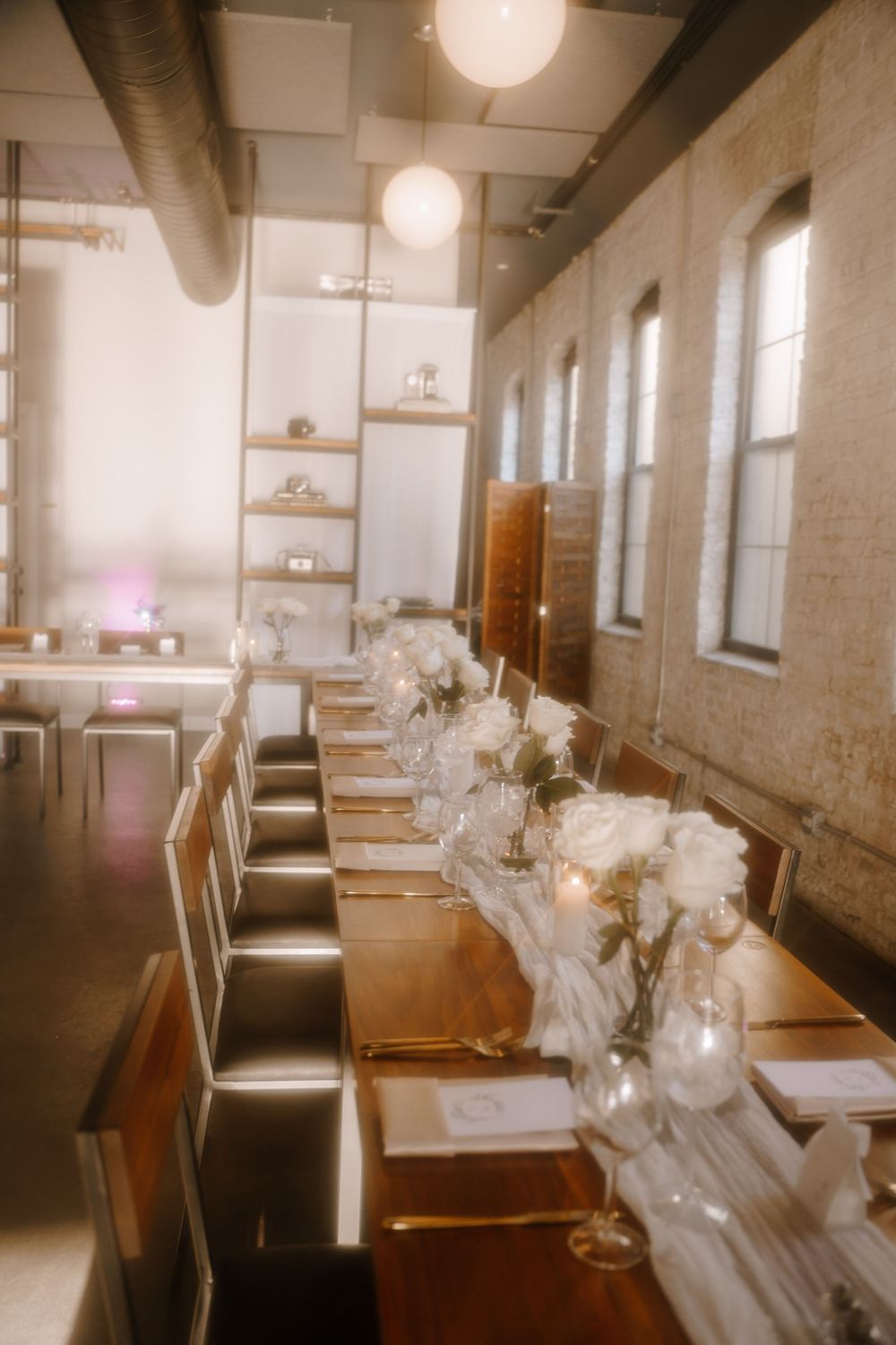 Long dining table set for an elegant event with white floral centerpieces in a rustic industrial space with tall windows.