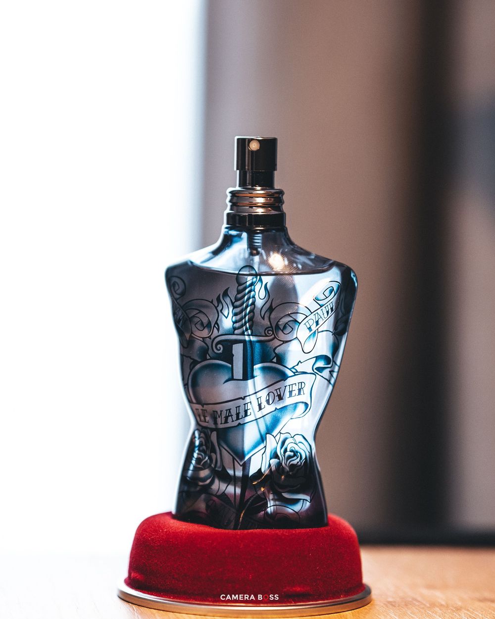 A blue glass perfume bottle with heart and tattoo designs sits on a red velvet display stand.