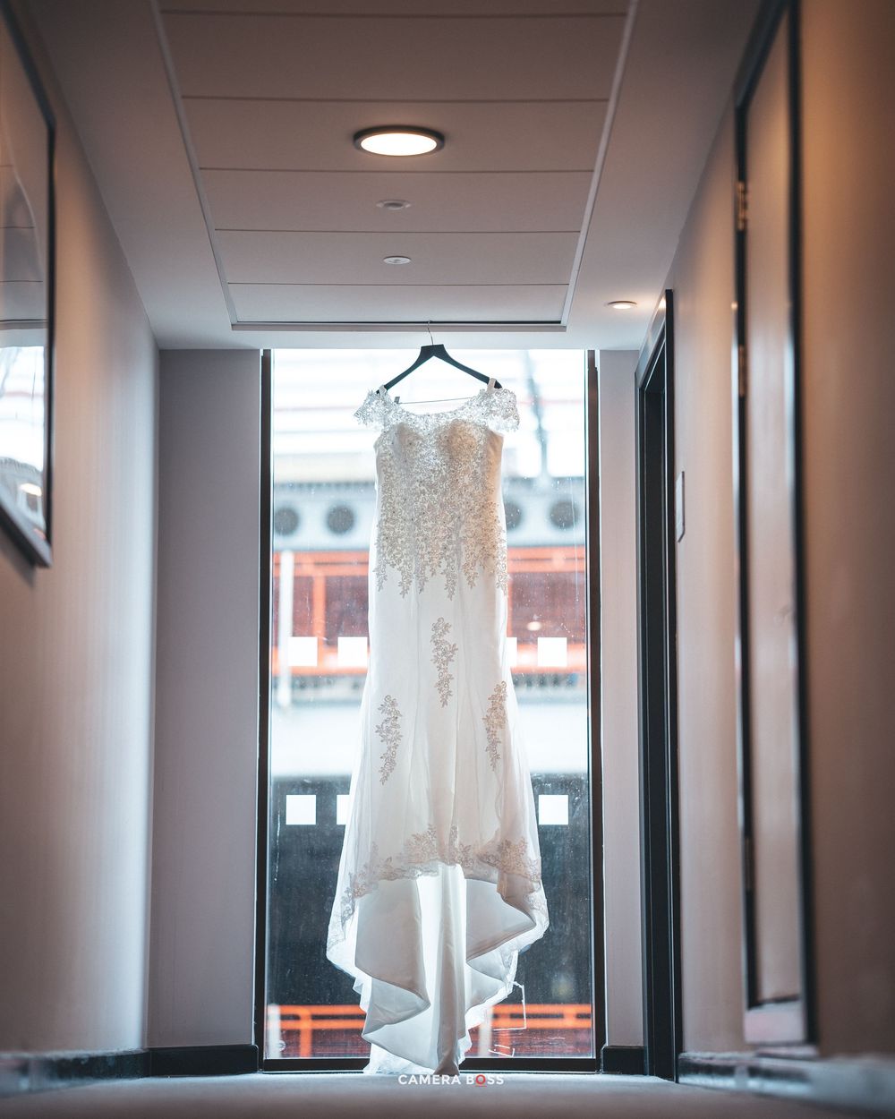 A white lace wedding dress hangs in silhouette against a window in a narrow hallway with recessed lighting.
