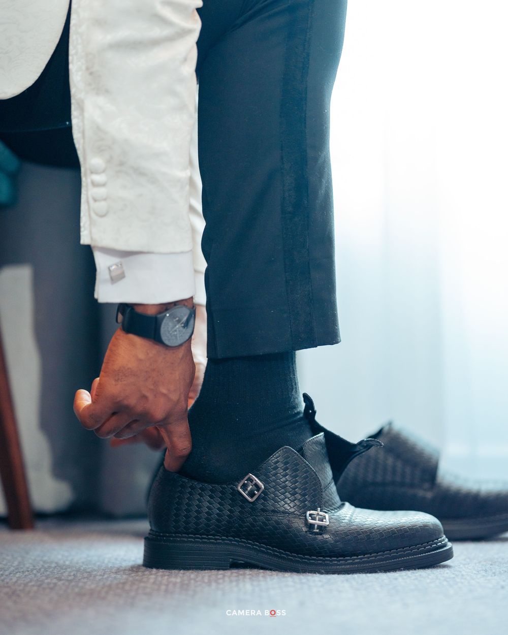Close-up fashion shot of a black leather monk strap dress shoe with textured pattern detail and cuffed light pants.
