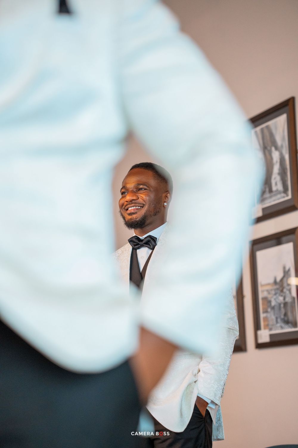 A side view through foreground objects captures someone in formal attire smiling warmly against a wall with framed artwork.