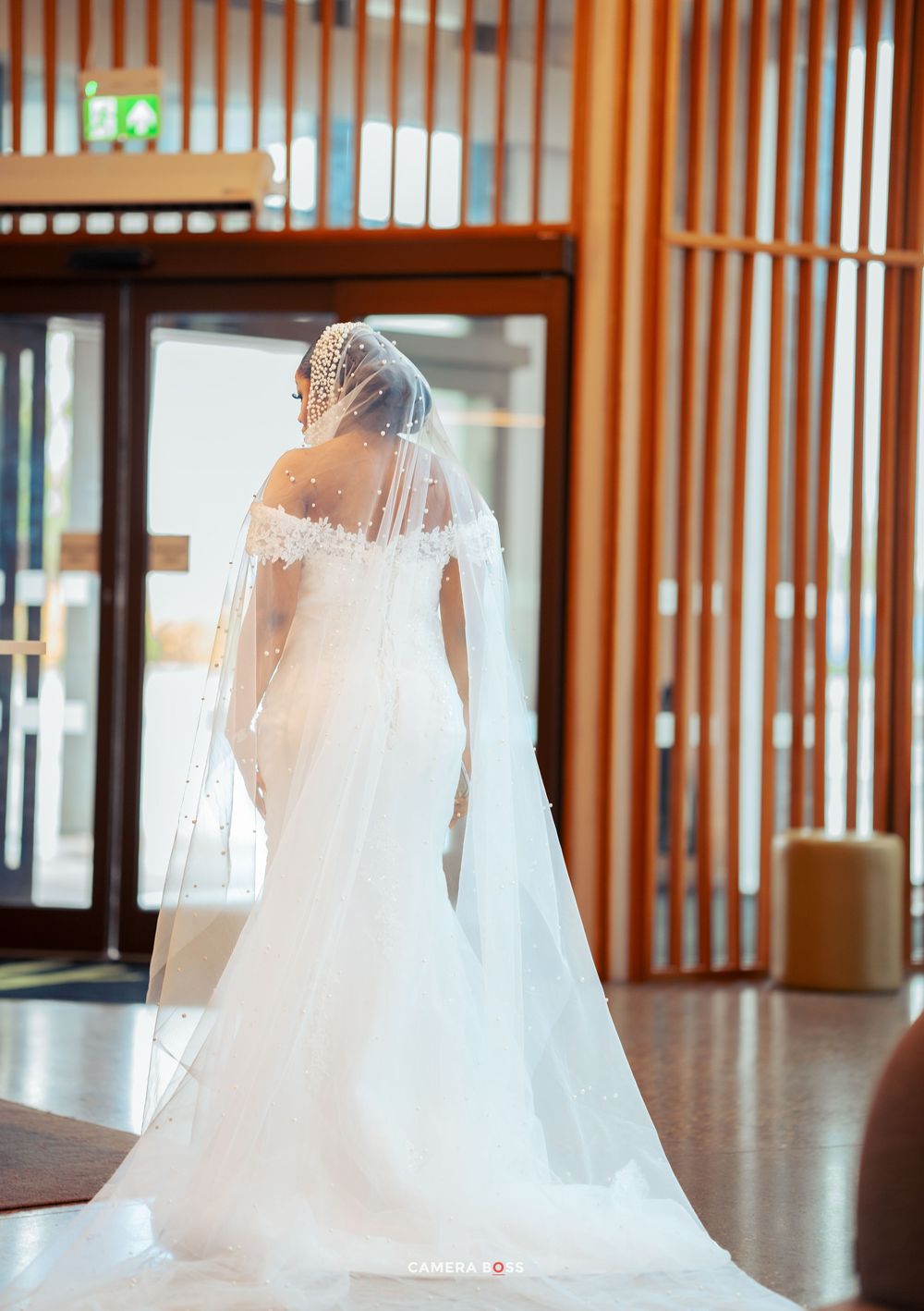 Elegant white wedding gown with flowing floor-length veil shown from behind in a modern venue with wooden accents.
