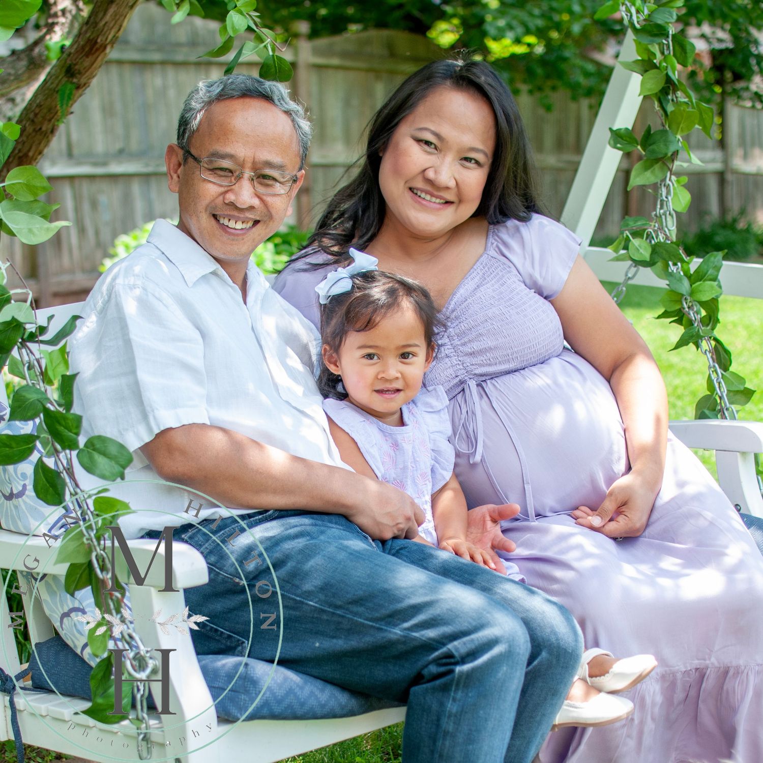 Mom, dad, and toddler sit together on a garden swing surrounded by green vines and leaves on a sunny day.