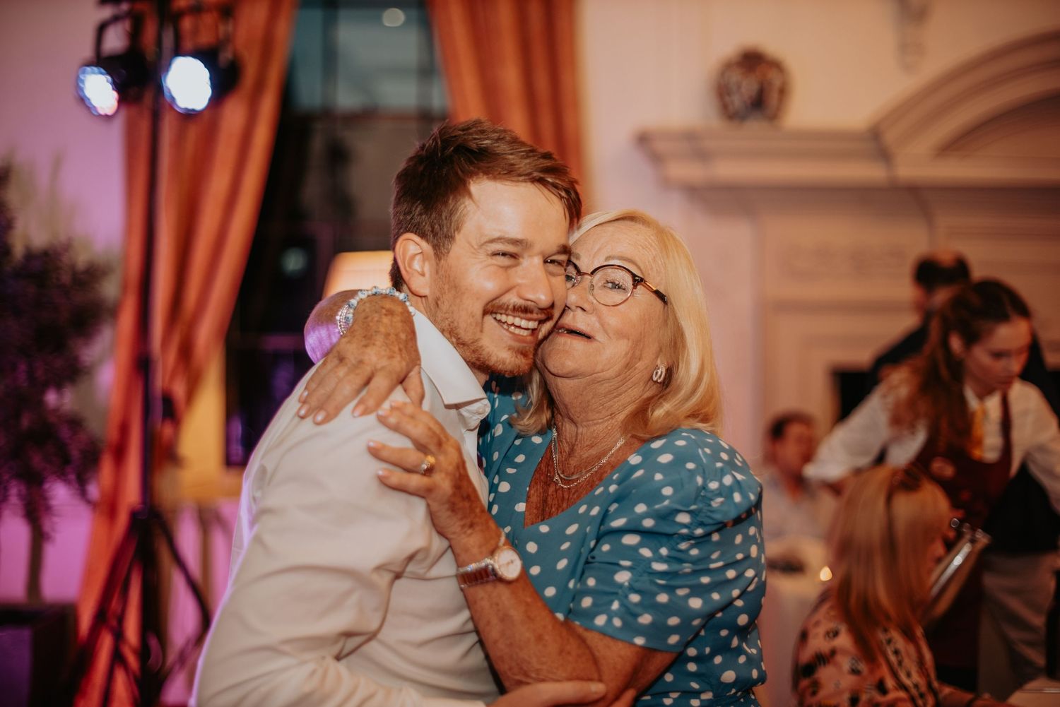 Two people share a warm embrace while dancing at an indoor event with soft pink lighting and decorative curtains in the background.