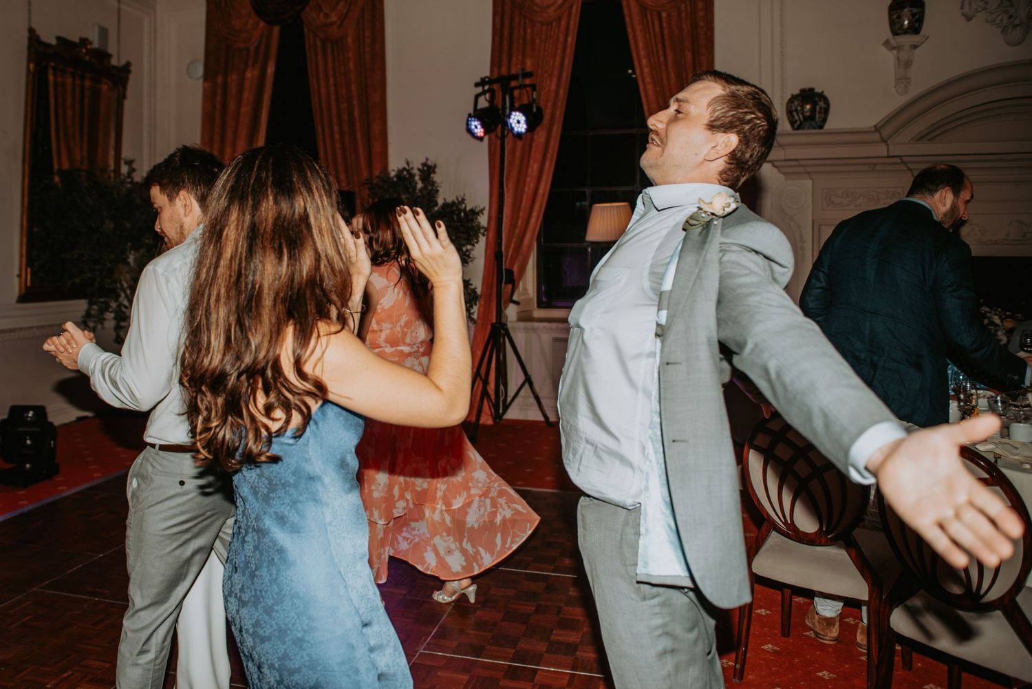 Party guests dance energetically in a vintage-style room with red curtains and decorative walls.