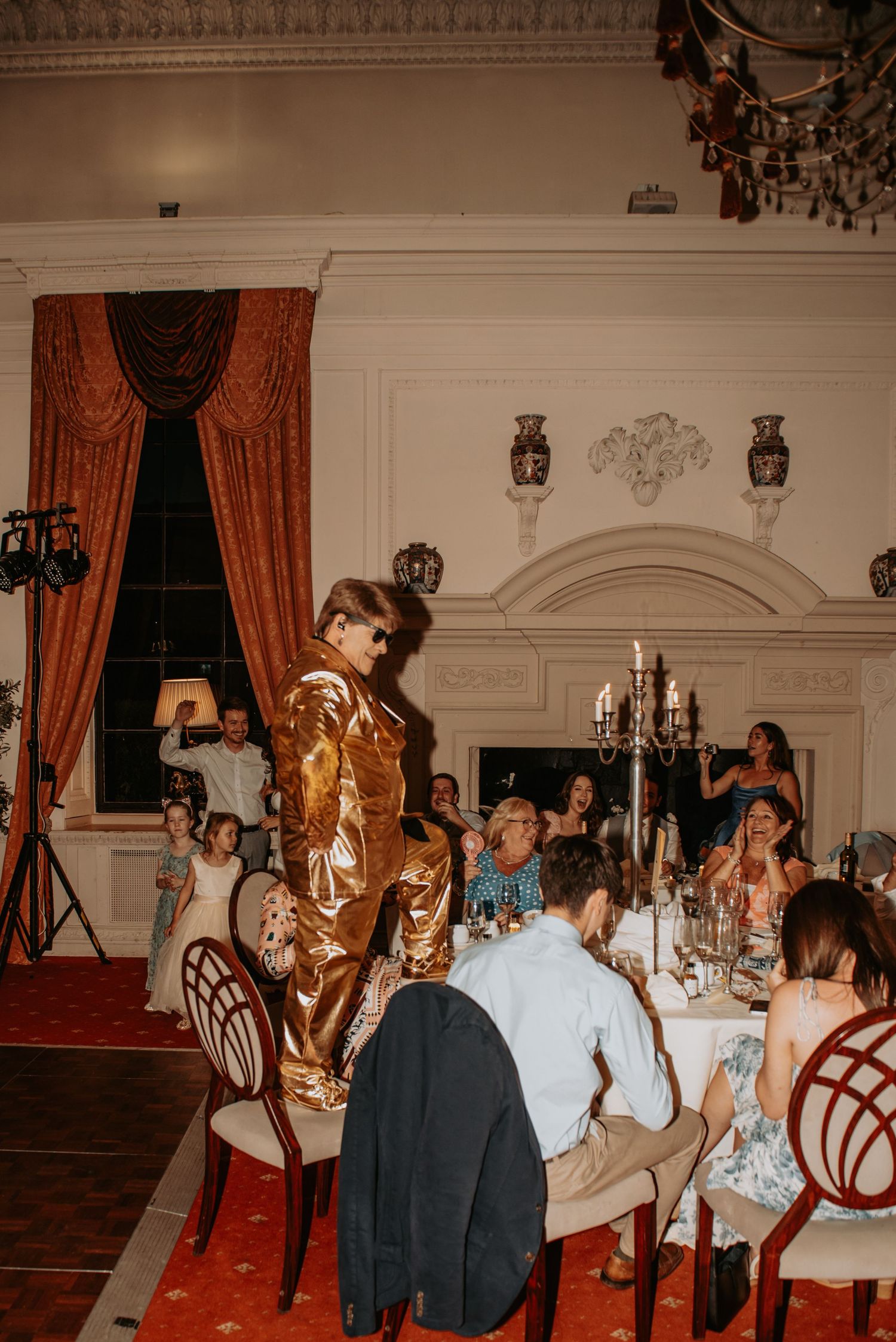 Elegant dinner party scene with candelabras and coral curtains in a vintage ballroom with ornate white walls and decor.