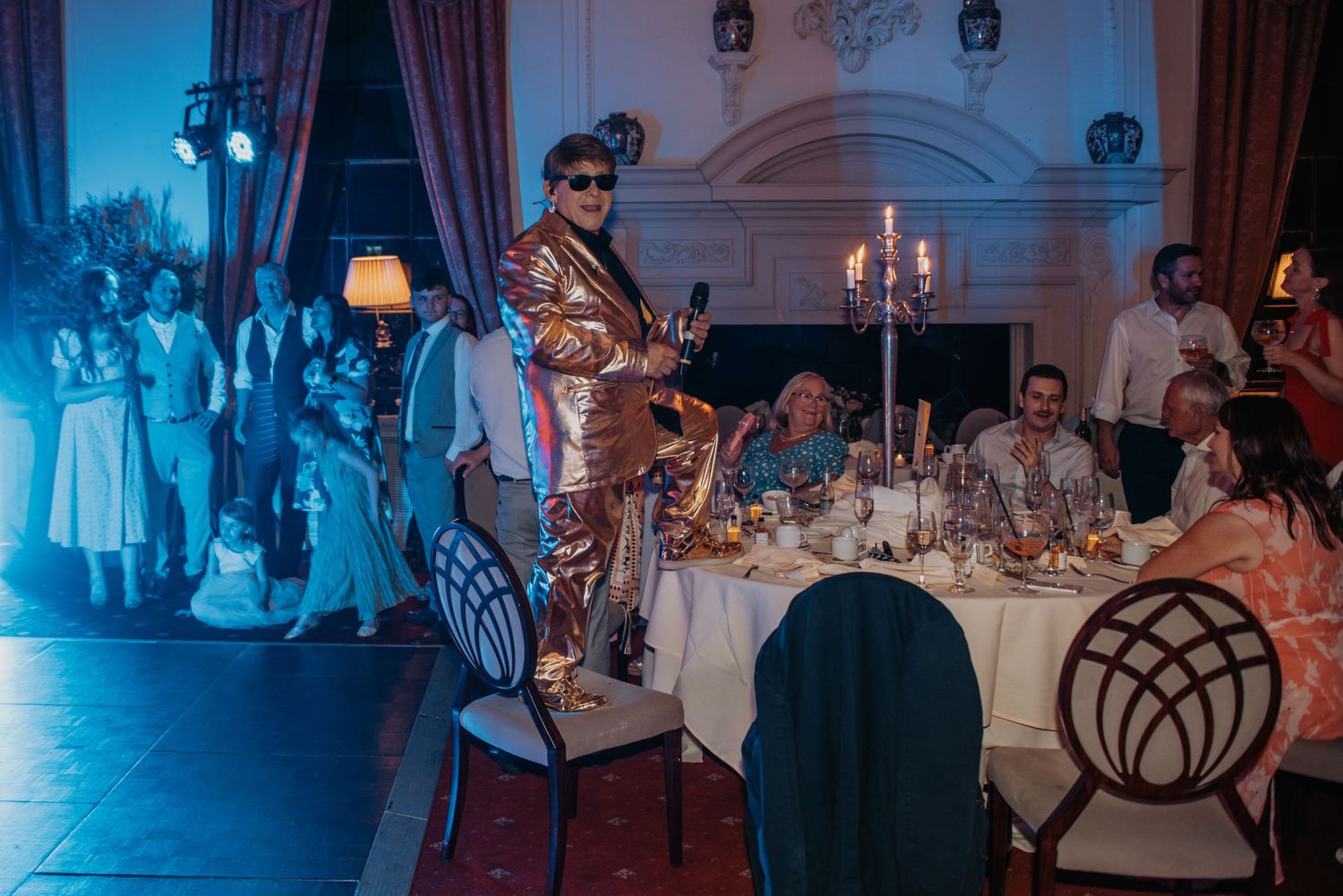 A performer in an ornate brown suit stands atop a chair during an elegant indoor dinner reception with candlelight.