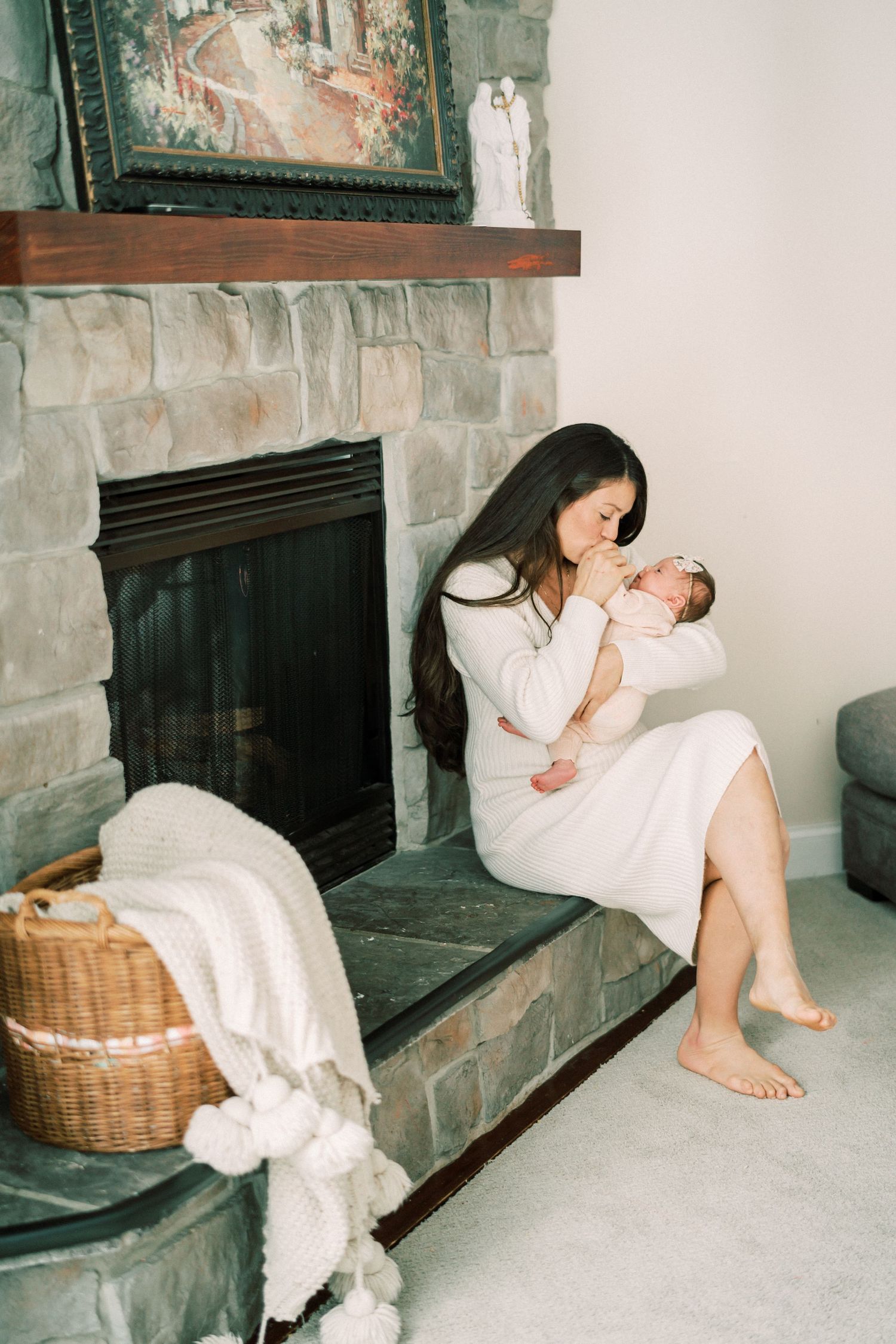 Mother in white dress sits on stone fireplace hearth while holding newborn.