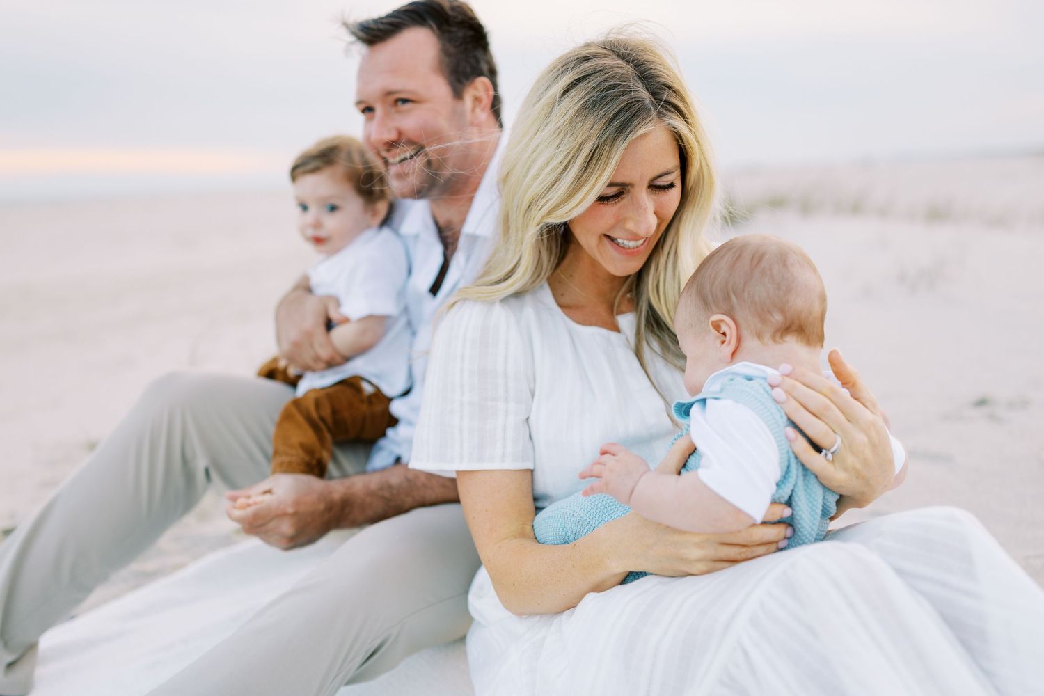 A family in white outfits sits together on a sandy beach during sunset, sharing a tender moment.