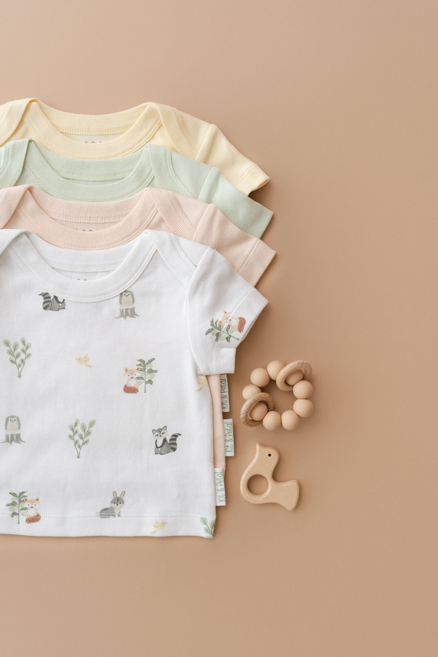 Baby onesies in pastel colors laid flat with wooden teething toys on beige background.