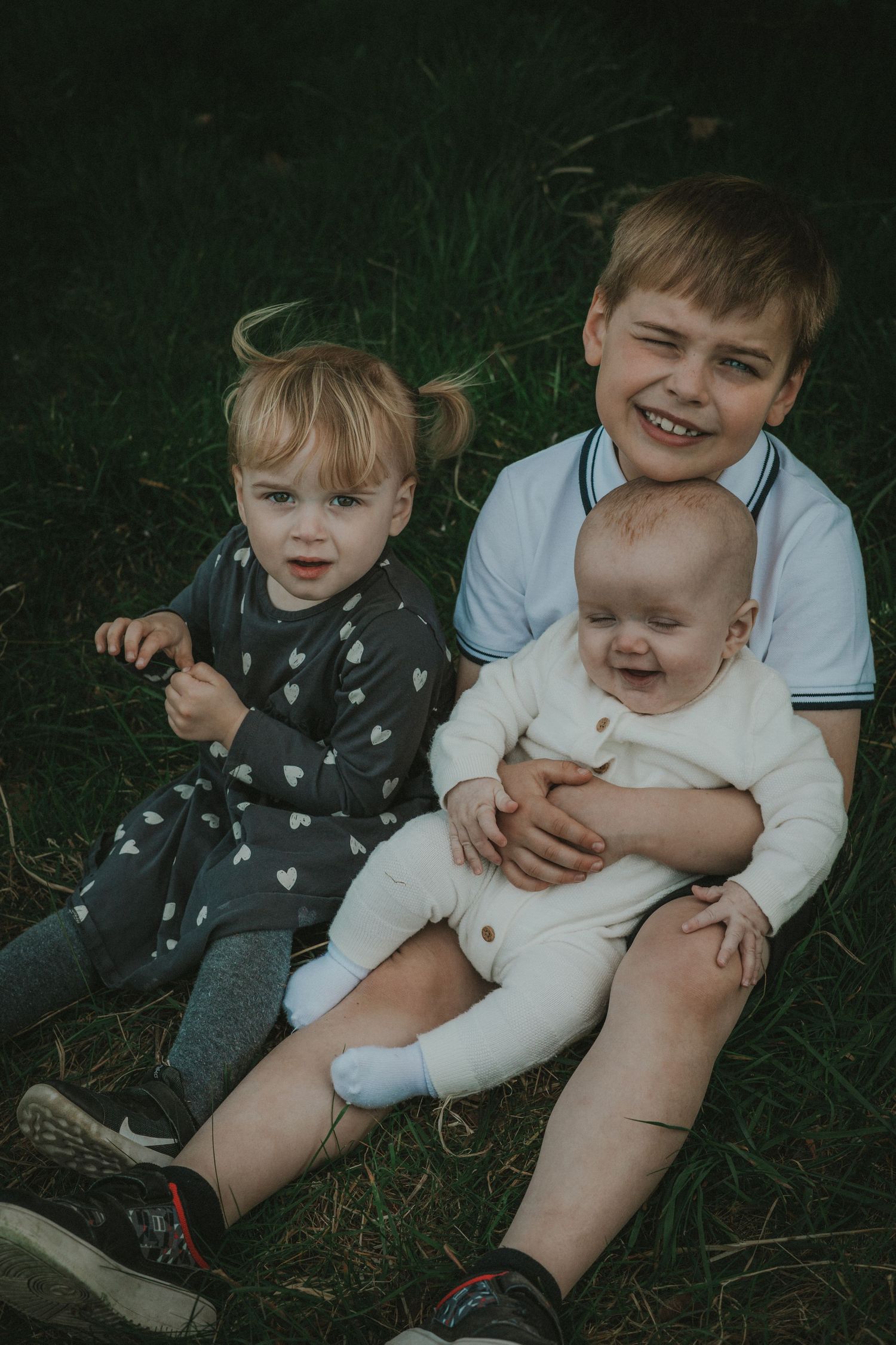 family session at needham market, three siblings, eldest holding youngest, sat in grass