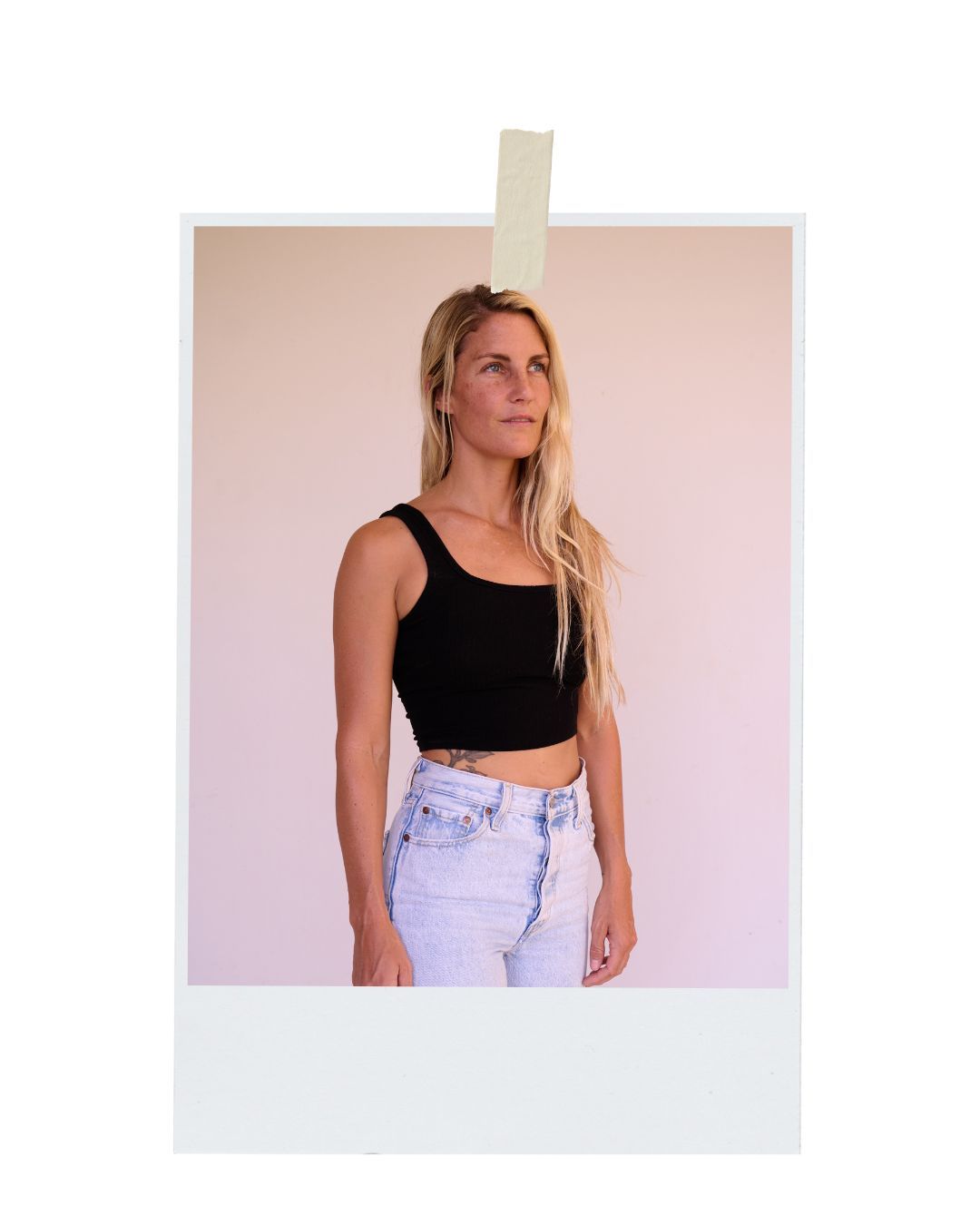 A person in a black crop top and light wash jeans poses against a pink backdrop in a polaroid-style photo.