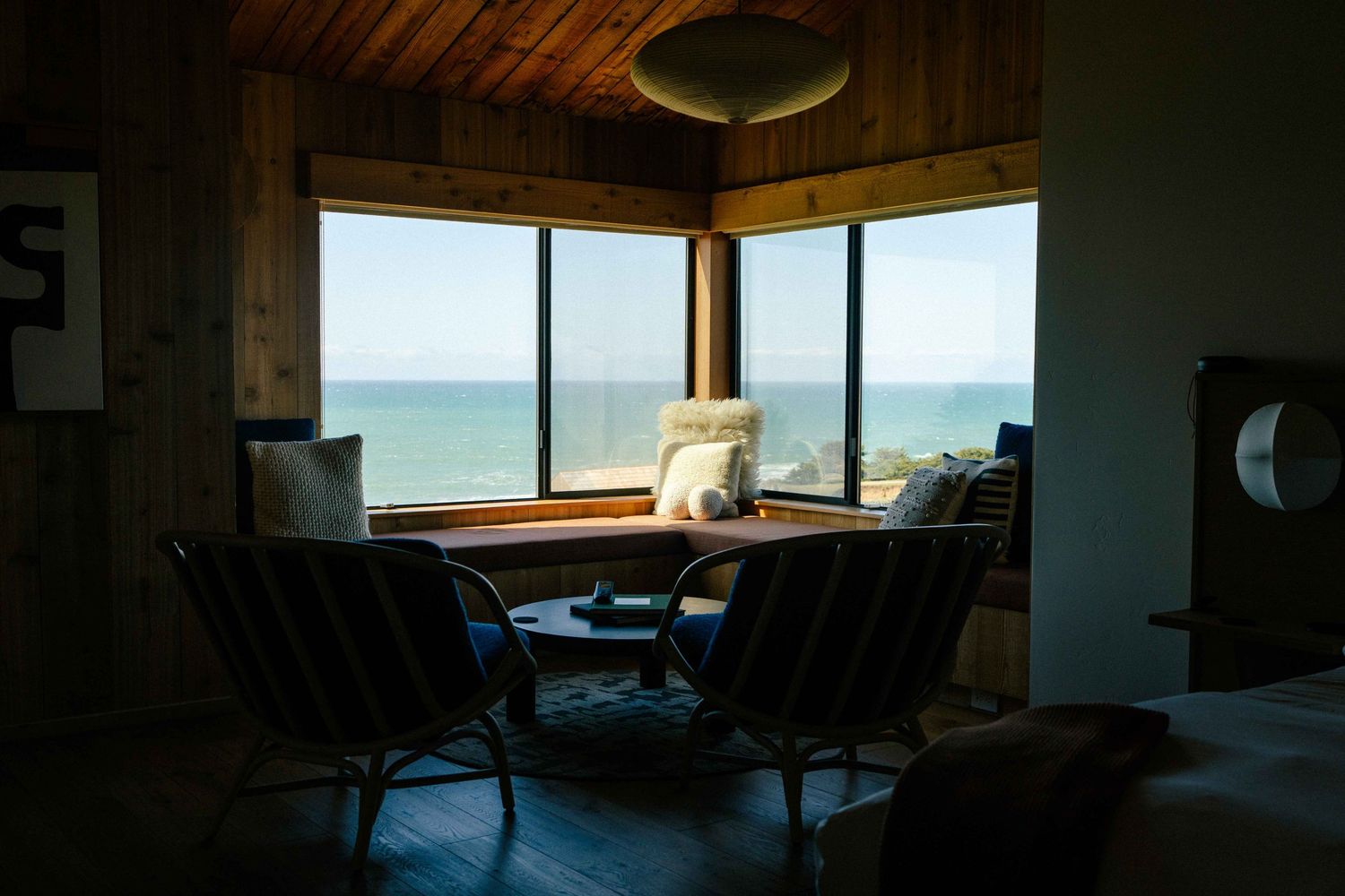 Dark interior room with corner windows overlooking the Pacific Ocean, featuring two wicker chairs at a wooden table.