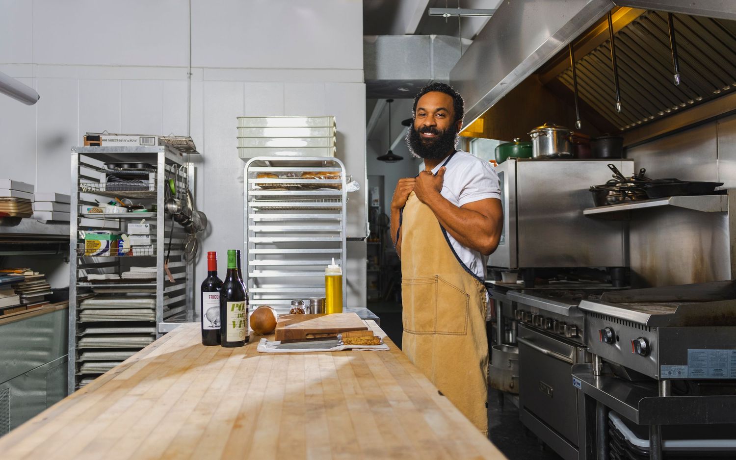 Restaurant worker in brown apron stands at wooden prep counter in commercial kitchen with stainless steel equipment.