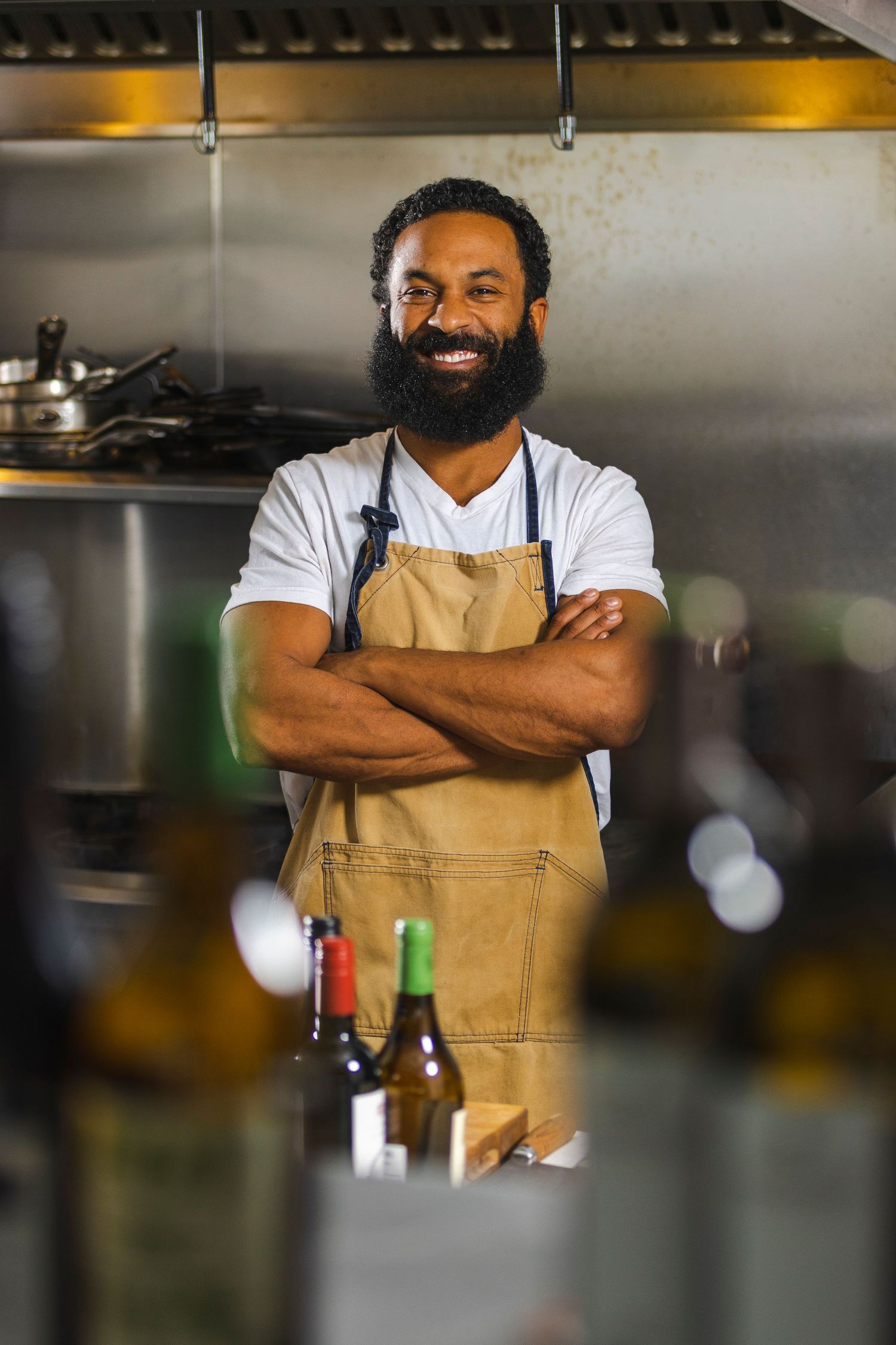 A smiling professional wearing a tan apron and white t-shirt stands confidently with crossed arms in a restaurant kitchen.