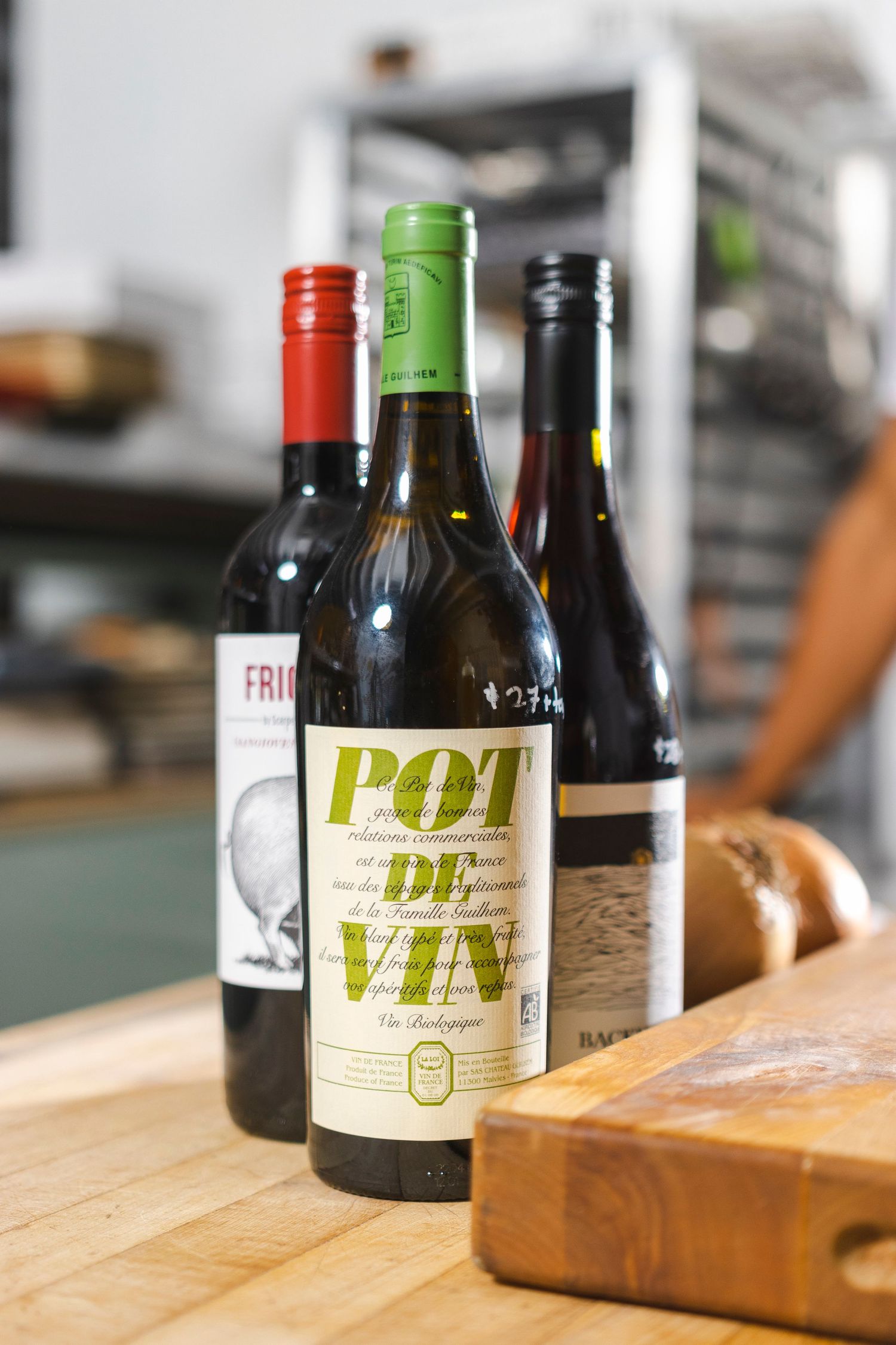 Three wine bottles including a 'Pot de Vin' bottle stand on a wooden cutting board against a blurred kitchen background.