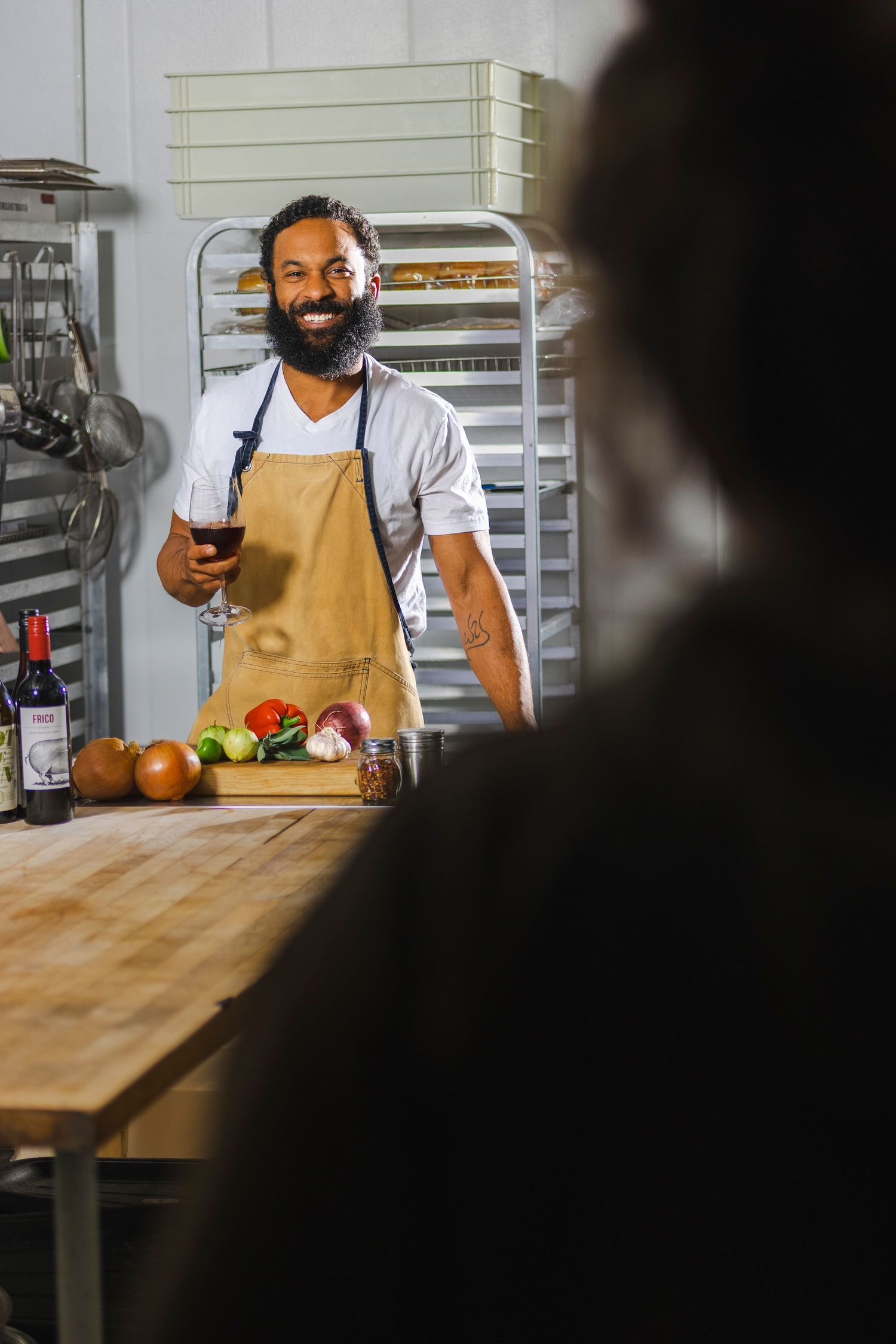 Chef in tan apron standing at wooden prep table with fresh produce and wine in commercial kitchen setting.