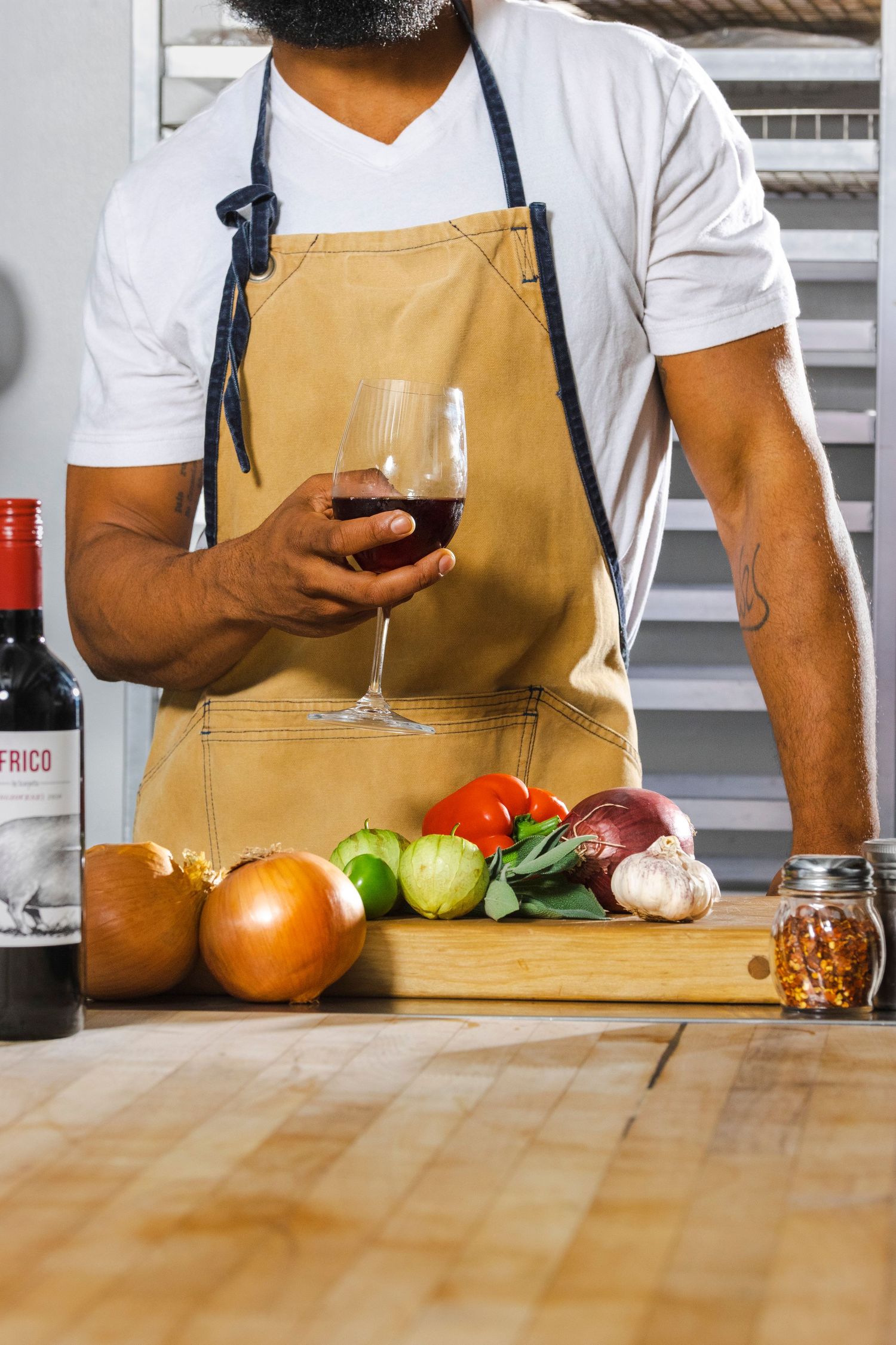 Chef wearing tan apron holding wine glass next to fresh vegetables and spices on wooden cutting board.