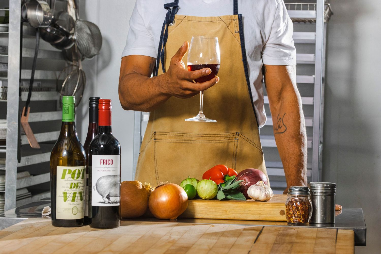 Person in tan apron holding wine glass with cooking ingredients and wine bottles on wooden cutting board.