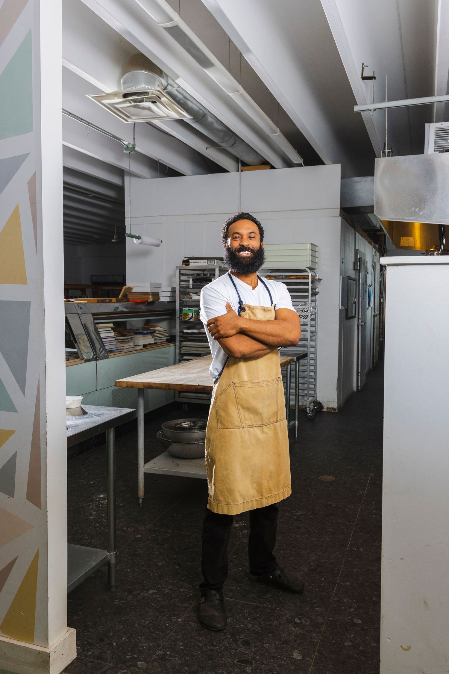 A professional wearing an apron and white shirt stands confidently in a commercial kitchen setting with exposed ceiling beams.