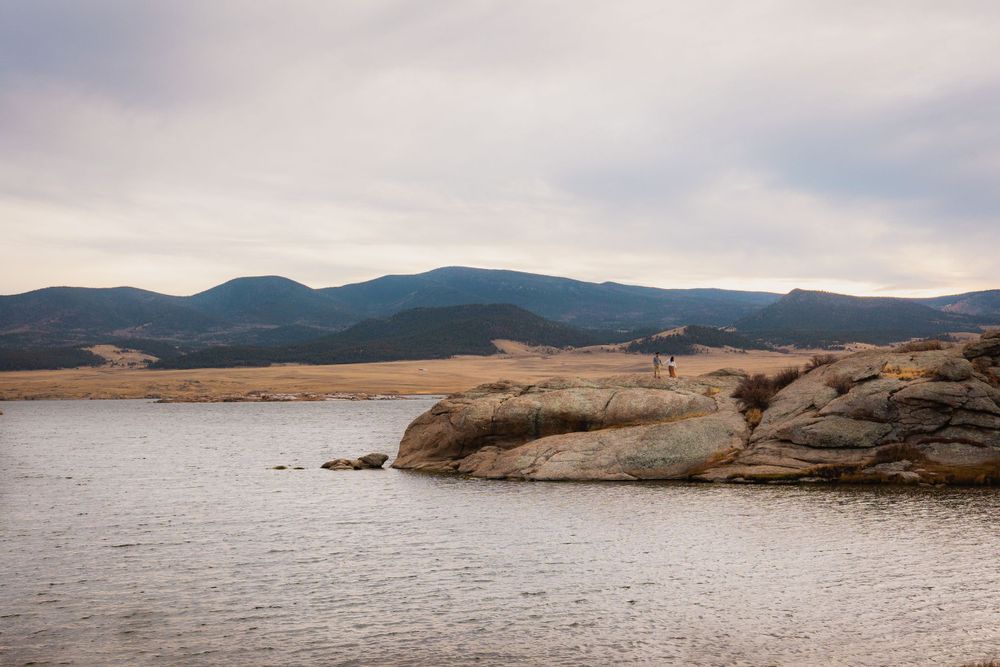 Rocky outcrop meets calm lake waters with mountains and cloudy skies in the background.