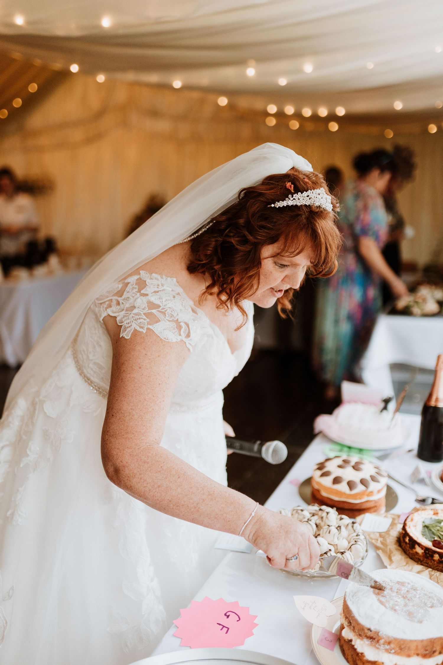 A joyful wedding reception moment as the bride in white lace dress and veil cuts into a layered naked cake.