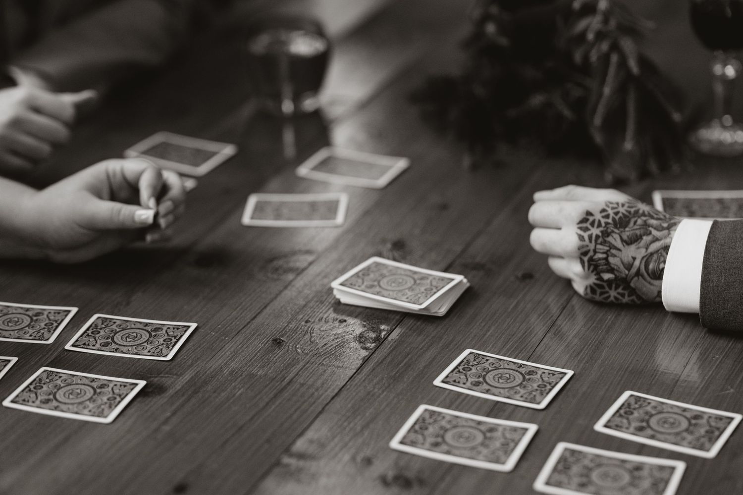 Black and white photograph showing hands around a wooden table playing a card game with scattered playing cards.
