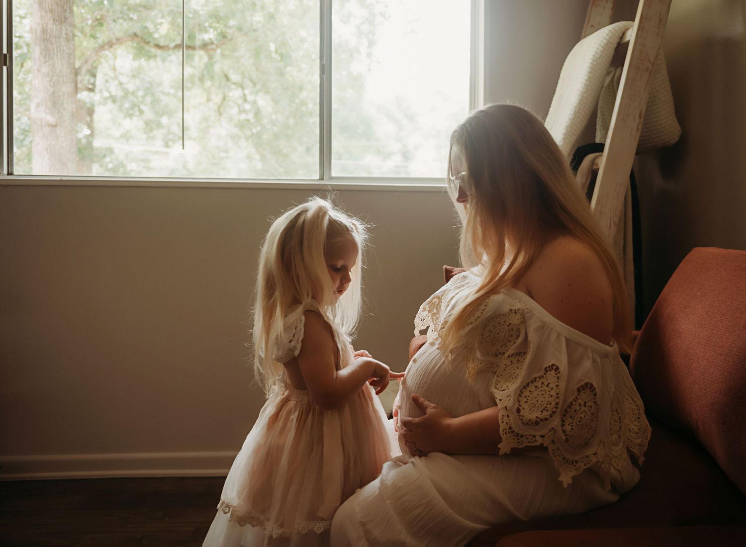 A tender moment between two figures in white dresses sitting by a sunlit window in a dreamy atmosphere.