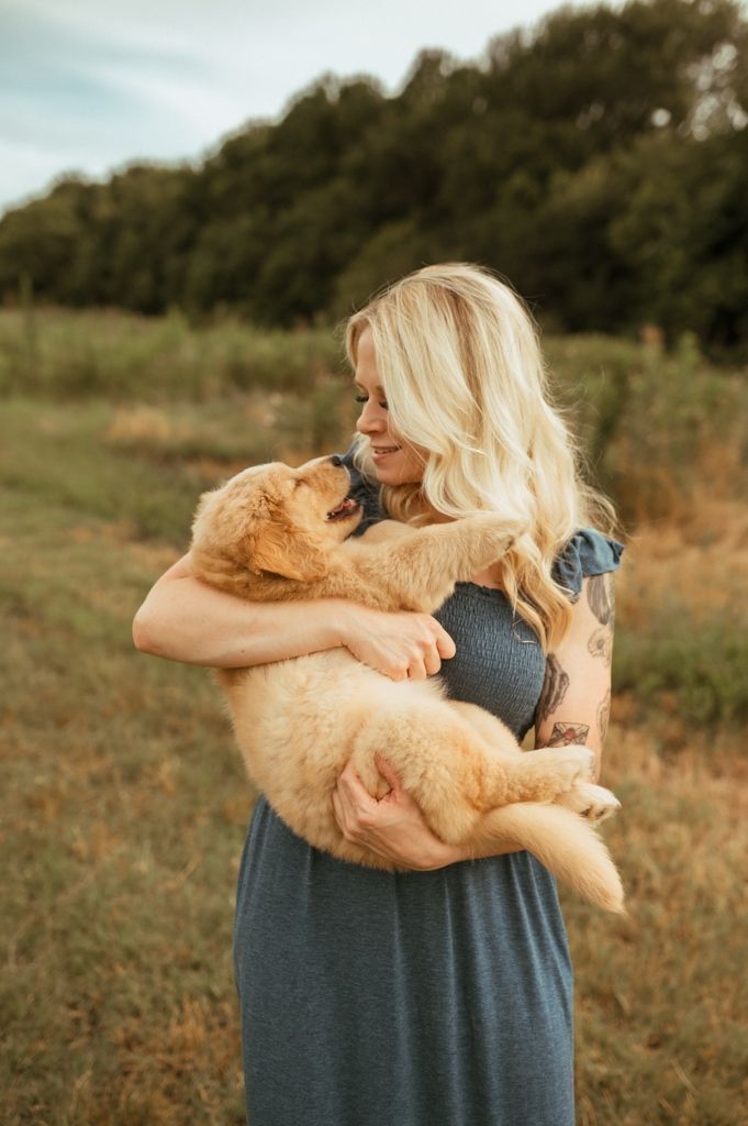 Golden retriever puppy being cuddled and held in arms while standing in grassy field.
