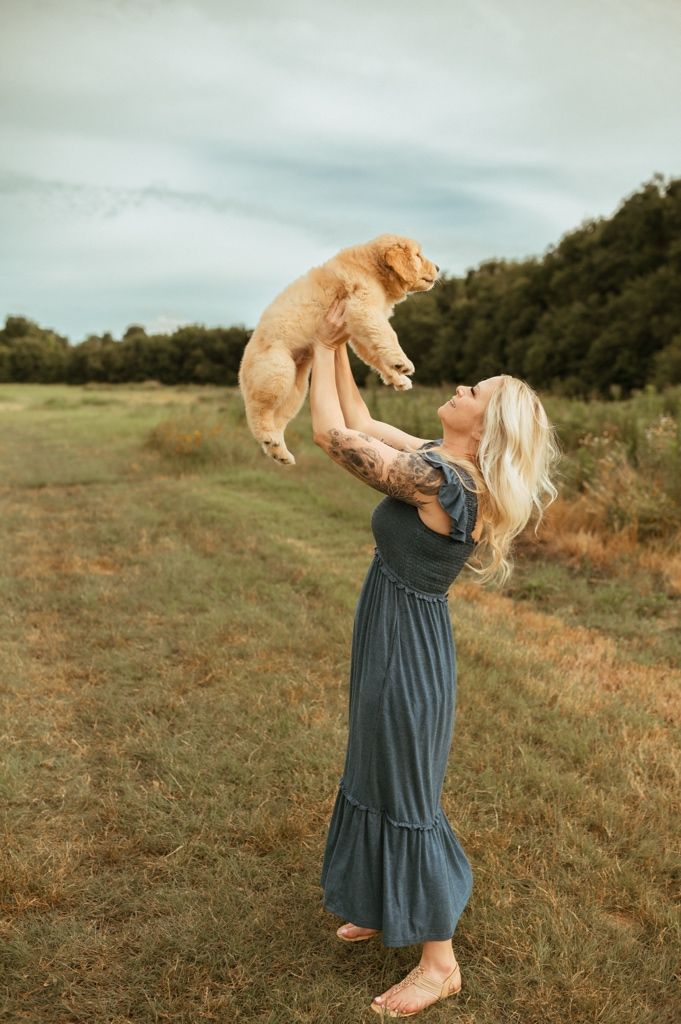 A Golden Retriever puppy is lifted playfully into the air in a grassy field on a cloudy day.