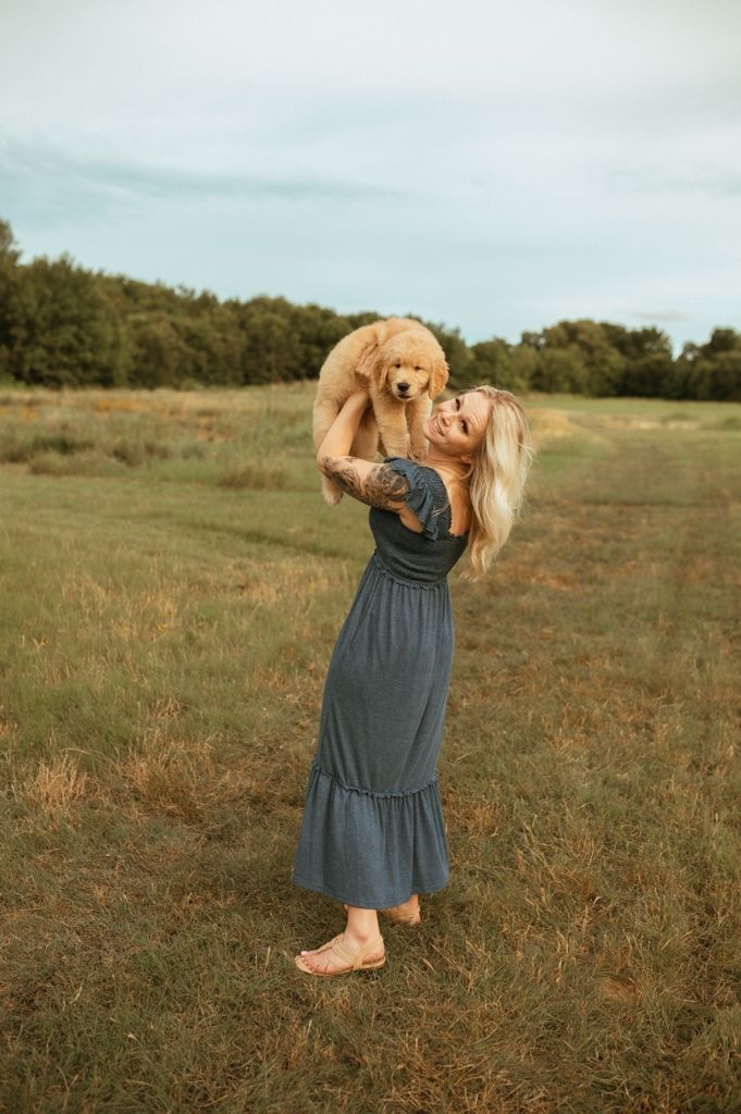 Golden retriever puppy held up high in blue dress against natural outdoor setting.