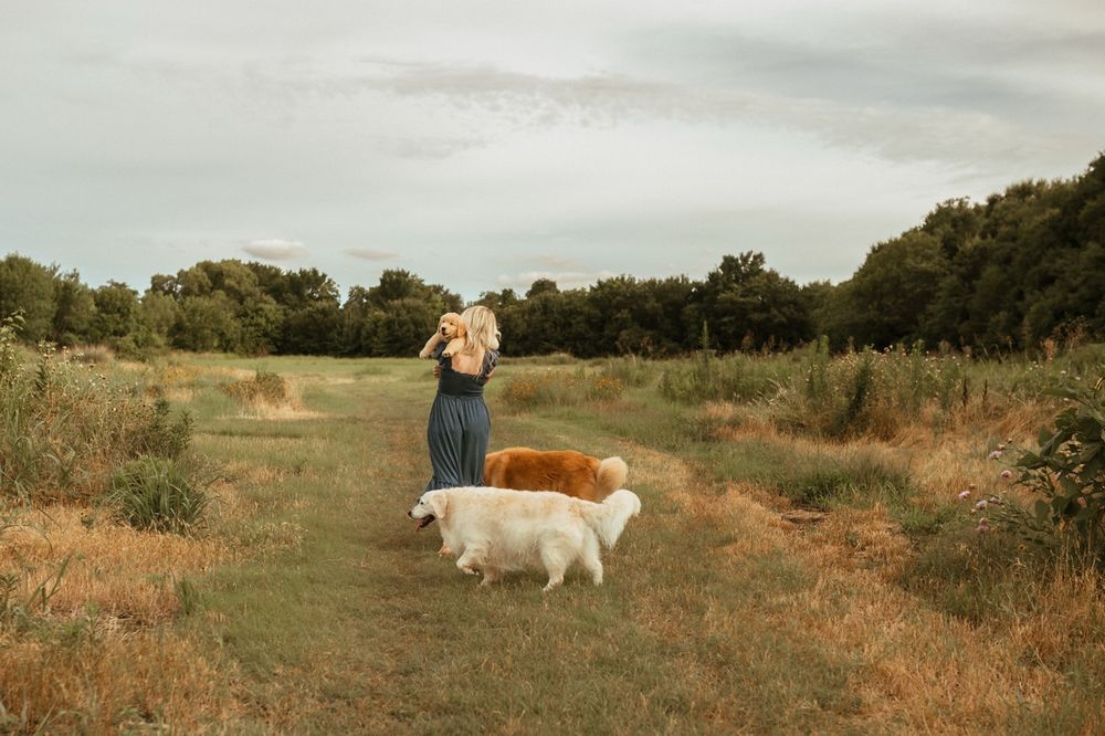 Someone in a flowy dress stands on a dirt path in a grassy field with two white dogs on a sunny autumn day.