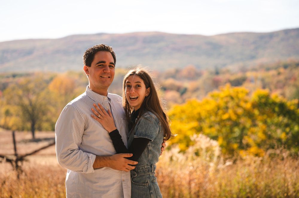 A couple embraces while posing outdoors against a scenic autumn mountain landscape with golden foliage in the background.
