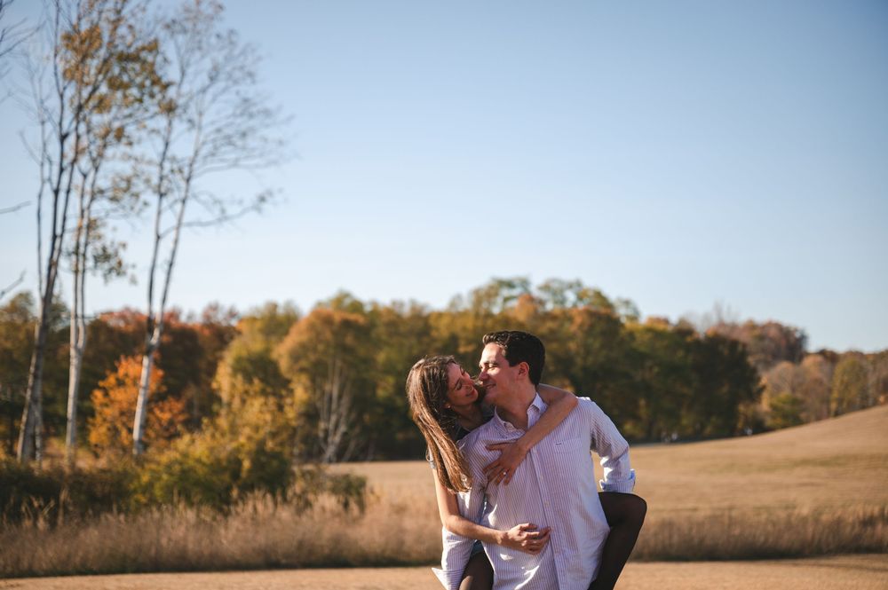 A couple embraces in a serene outdoor setting with golden fall trees and fields in the background.