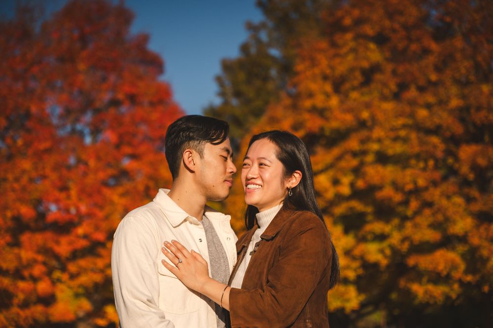 A loving couple embraces outdoors surrounded by vibrant autumn foliage with red and orange maple trees.